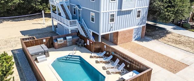 Elevated blue and white beach house with pool, lounge chairs, and deck surrounded by sand and vegetation