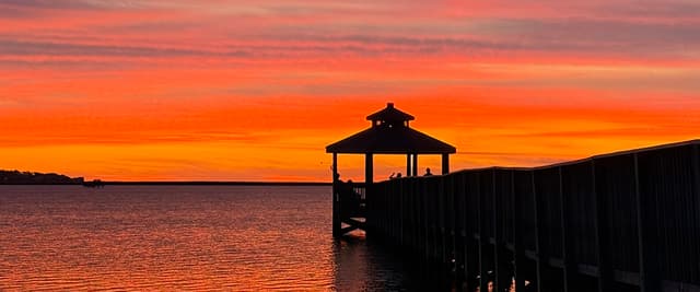 Silhouetted gazebo at end of wooden pier at sunset over calm water