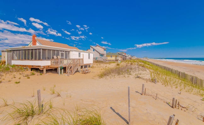 Beachfront houses on stilts with dunes, beach grass, and wooden fence along sandy shore under blue sky