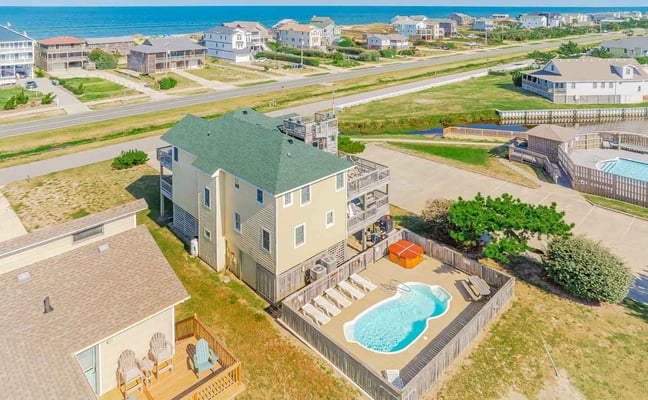 Aerial view of yellow beachfront house with green roof, private pool, and deck overlooking ocean and neighboring coastal homes