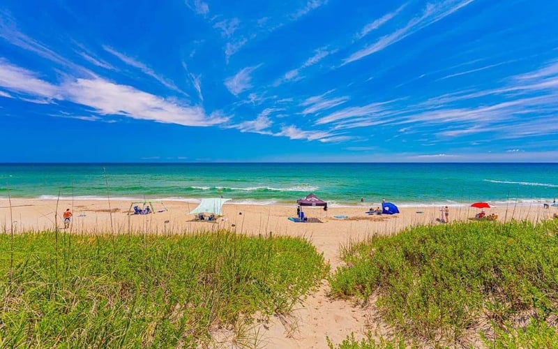 Sandy beach with dune grass, colorful umbrellas, and swimmers in turquoise ocean under clear blue sky